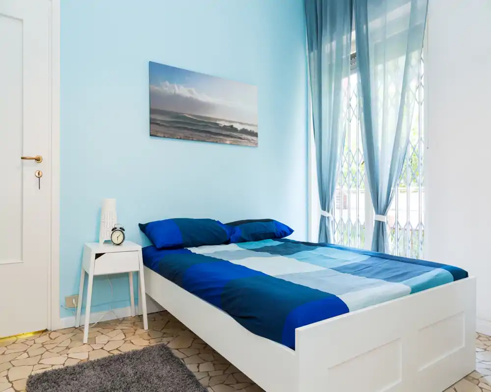 Camera da letto luminosa con letto matrimoniale ben rifatto, pareti azzurre e tende leggere che lasciano filtrare la luce naturale — perfetta per un soggiorno rilassante.