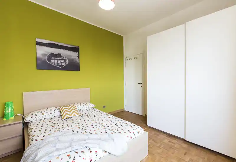 Camera da letto moderna e luminosa con letto matrimoniale, biancheria decorata, comodino e un ampio armadio bianco su una parete verde.
