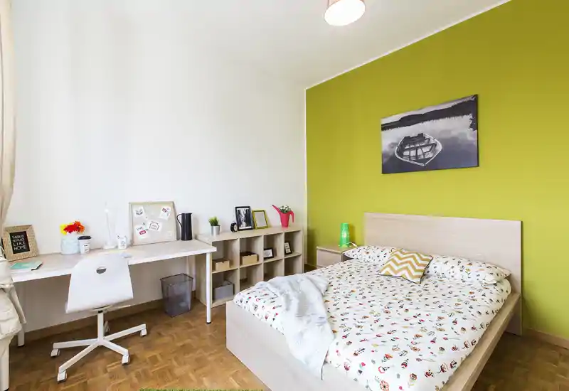 Camera da letto luminosa e moderna con letto matrimoniale, biancheria a fantasia, comodino e scrivania ordinata davanti a una parete accentata verde lime.