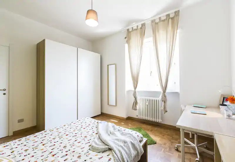 Camera da letto luminosa e minimal con letto matrimoniale, ampia armadiatura, scrivania e finestra con tende leggere che lasciano entrare la luce naturale.