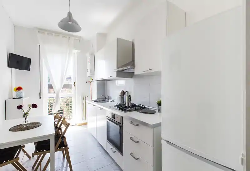 Cucina moderna e luminosa con mobili bianchi, piano cottura a gas, forno integrato e un piccolo tavolo da pranzo vicino alla porta che dà sul balcone — perfetta per appartamenti in città.