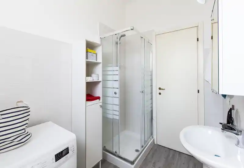 Bagno moderno e luminoso con box doccia angolare in vetro, lavabo bianco e mobiletto contenitore. Piastrelle chiare e pavimento effetto legno per un aspetto pulito e fresco.