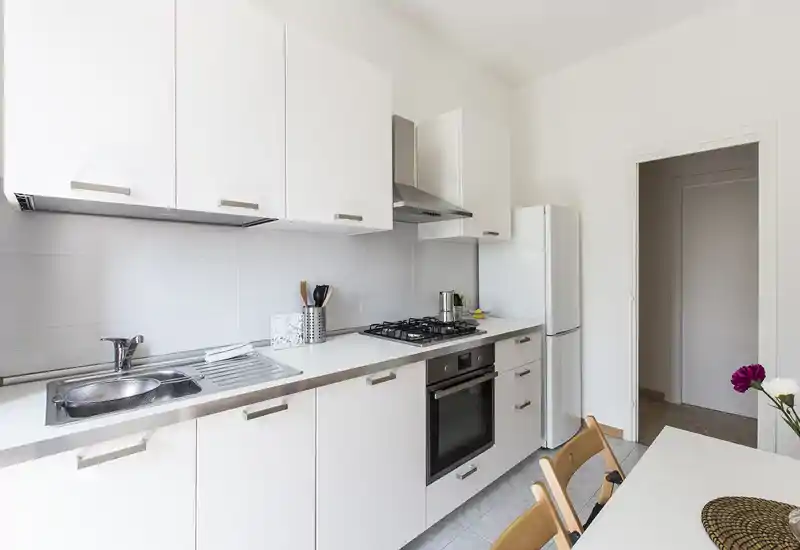 Cucina moderna e luminosa con pensili bianchi, lavello in acciaio, piano cottura a gas e forno incassato; piccolo angolo pranzo visibile. Design pulito e minimale perfetto per annunci di affitto.