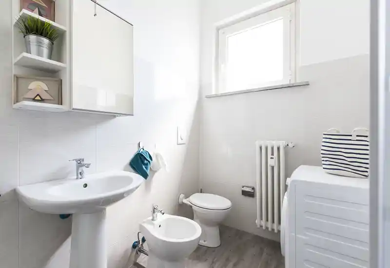 Bagno moderno e luminoso con lavabo a colonna, bidet, WC, termosifone e lavatrice; piastrelle neutre e una finestra alta assicurano un aspetto pulito e arioso.
