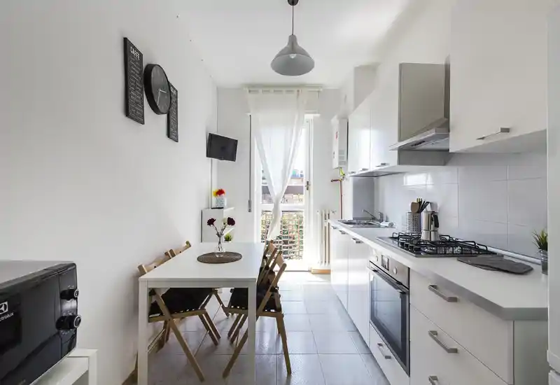 Cucina moderna e luminosa con piccolo tavolo da pranzo per quattro, mobili bianchi, forno integrato e piano cottura a gas, con luce naturale dalla porta del balcone — perfetta per pasti accoglienti.