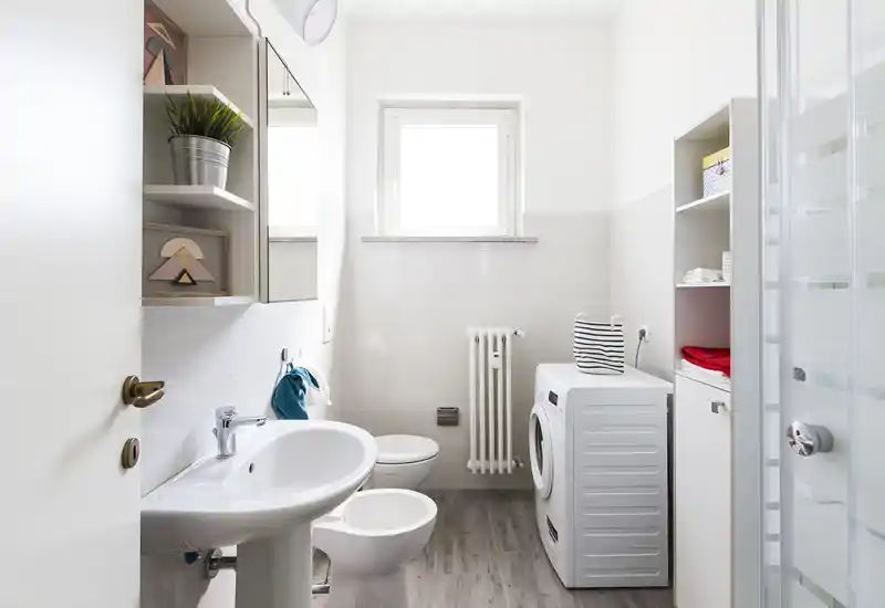 Bagno moderno e luminoso con lavabo, bidet, WC, box doccia e lavatrice; piastrelle neutre e luce naturale da una finestra rendono l'ambiente pulito e fresco.