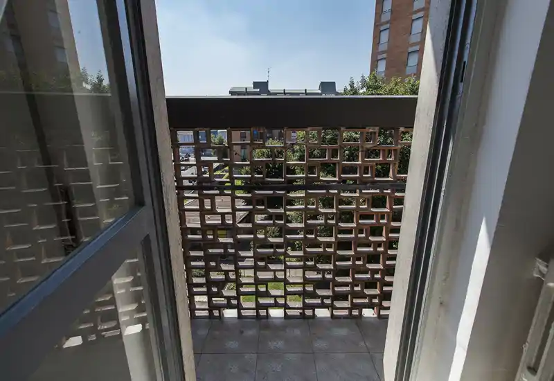 Piccolo balcone piastrellato con ringhiera in mattoni traforati, vista su un cortile e palazzine vicine; luce diurna intensa con ombre parziali.