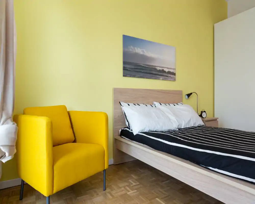 Camera da letto moderna e luminosa con letto su pedana in legno, biancheria a righe bianco-nera, lampada da comodino e una poltrona gialla come elemento d'accento. Pavimento in parquet e quadro con scena marina sopra la testiera.