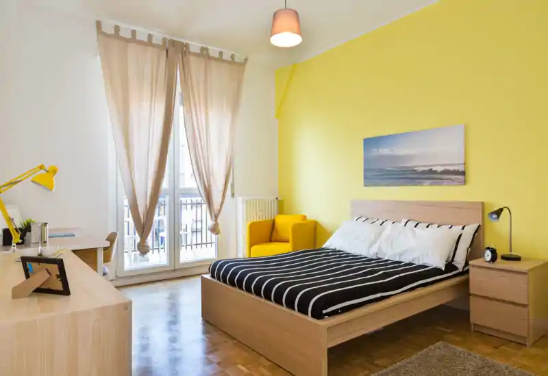 Camera da letto moderna e luminosa con letto matrimoniale davanti a una parete gialla, luce naturale dalle porte del balcone e una poltrona accogliente — perfetta per annunci in affitto.