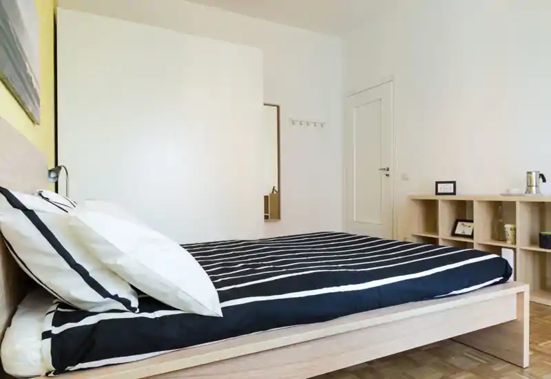 Camera da letto moderna e luminosa con letto basso in legno e biancheria a righe bianco e nero, cuscini morbidi e arredi minimal. Ampio armadio e mensole aperte offrono praticità senza appesantire l'ambiente.