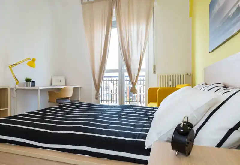 Camera da letto moderna e luminosa con un grande letto con biancheria a righe nere e bianche, una scrivania con lampada gialla e una poltrona gialla vicino alla finestra; ideale per soggiorni confortevoli e luminosi.