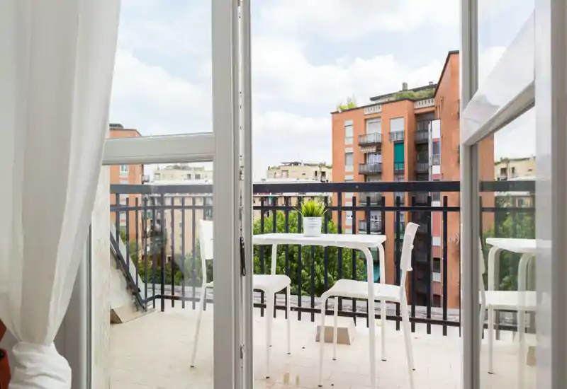 Piccolo balcone in città con tavolo e sedie bianche, vaso con pianta e vista sugli edifici in mattone — luminoso e ideale per il caffè del mattino.