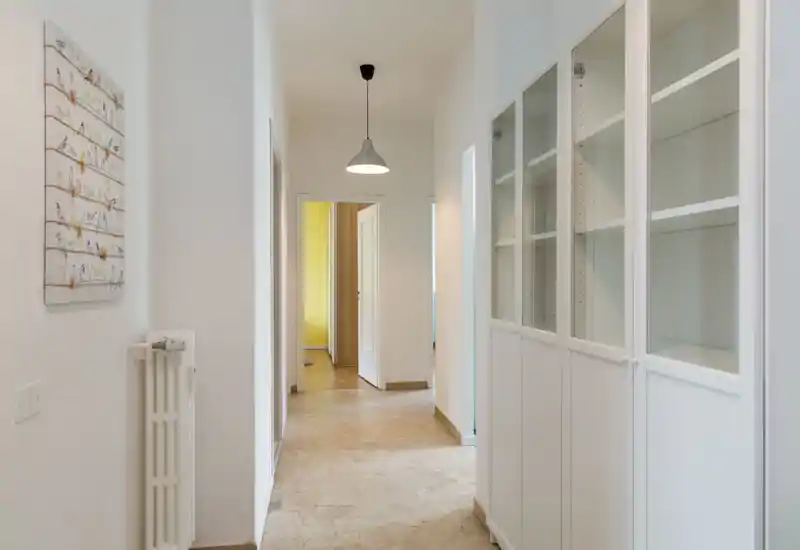 Ingresso/corridoio luminoso e pulito con armadi bianchi a parete e un lampadario a sospensione, che conduce alle stanze; pavimento in marmo neutro e pareti bianche creano una sensazione di spazio.