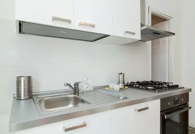 Cucina moderna e luminosa con lavello in acciaio inox, piano cottura a gas e forno integrato. Pensili bianchi e styling minimale creano uno spazio pulito e funzionale.
