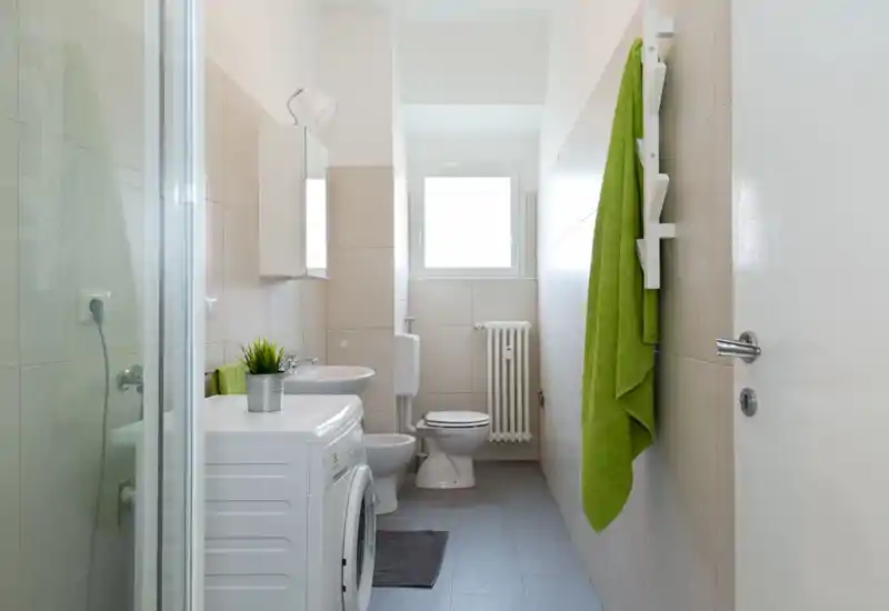 Bagno moderno e luminoso con wc, lavabo, doccia e lavatrice. Piastrelle neutre, termosifone sotto la finestra e asciugamani verdi a dare un tocco fresco.