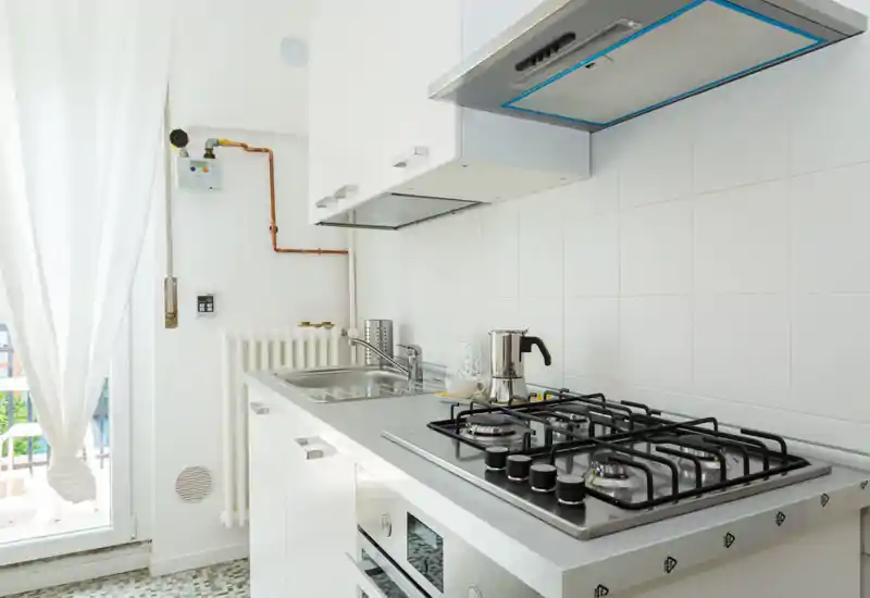 Cucina moderna e luminosa con piano cottura a gas in acciaio, cappa, lavello e mobili bianchi minimal. La luce naturale dalla porta crea un'atmosfera fresca e ordinata, perfetta per annunci.