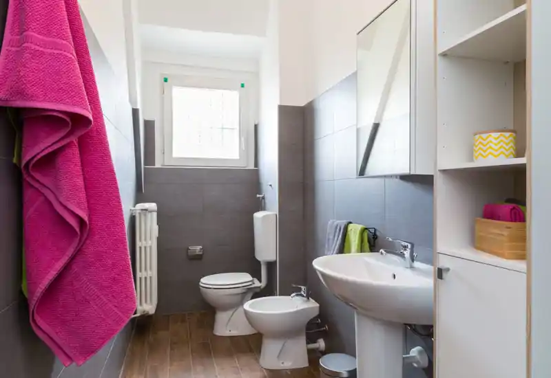 Bagno moderno e luminoso con lavabo, bidet e WC, piastrelle grigie e pavimento effetto legno; asciugamani colorati donano vivacità.