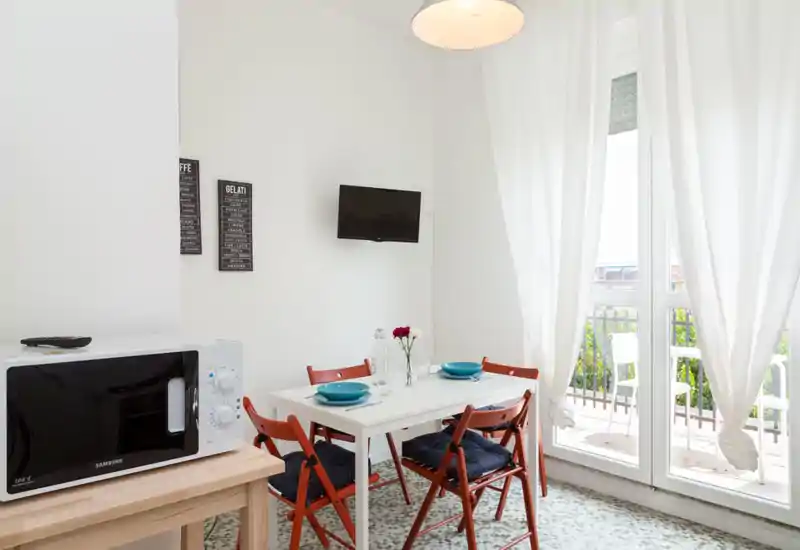Angolo pranzo luminoso e compatto con un piccolo tavolo bianco apparecchiato per due, sedie pieghevoli rosse con cuscini e grandi porte-finestre che si aprono su un balcone e illuminano l'ambiente.