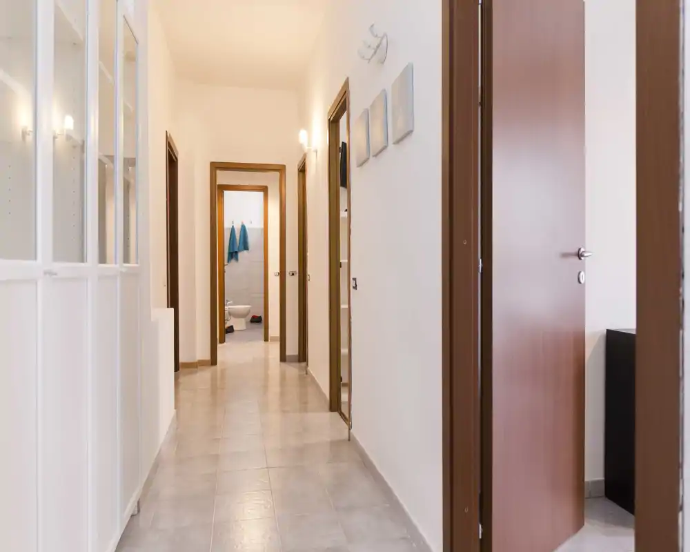 Corridoio/ingresso stretto con porte visibili e pavimento piastrellato; l'immagine è digitalmente distorta ma mostra un corridoio che conduce a più stanze.