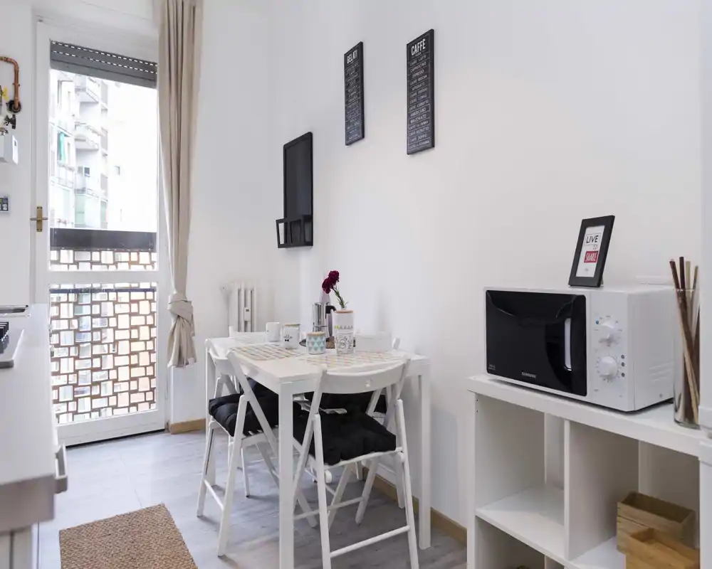Zona pranzo moderna e luminosa con un piccolo tavolo rotondo apparecchiato e sedie vicino a finestre alte—stile minimalista e luce naturale.