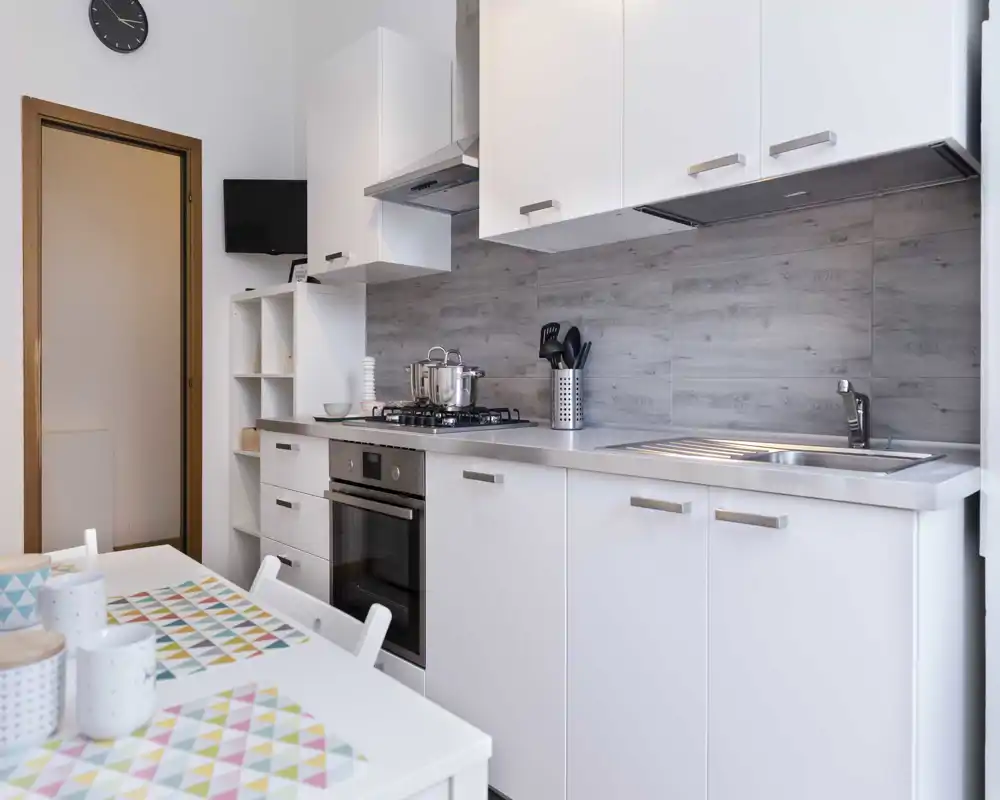 Cucina moderna e compatta con mobili bianchi, piccoli elettrodomestici sul piano e paraschizzi piastrellato. Spazio luminoso e contemporaneo, adatto per affitti brevi.