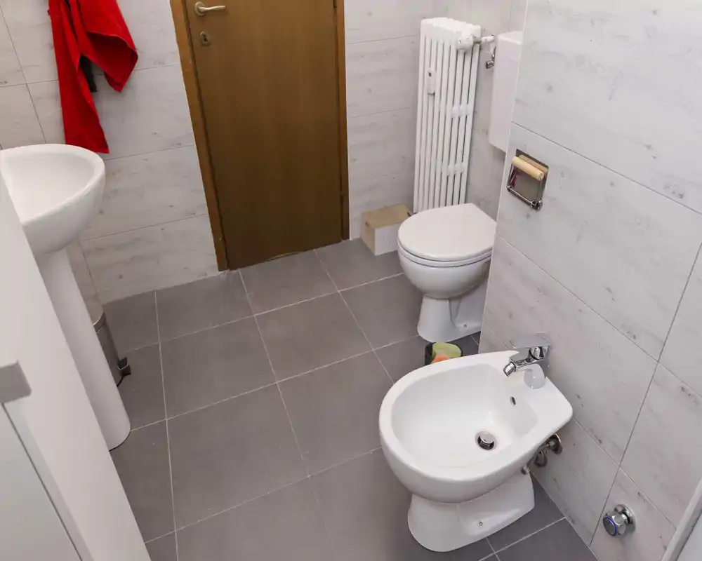 Bagno compatto con WC e lavabo a muro; visibili piastrelle neutre e un radiatore verticale. La foto mostra un piccolo bagno funzionale.
