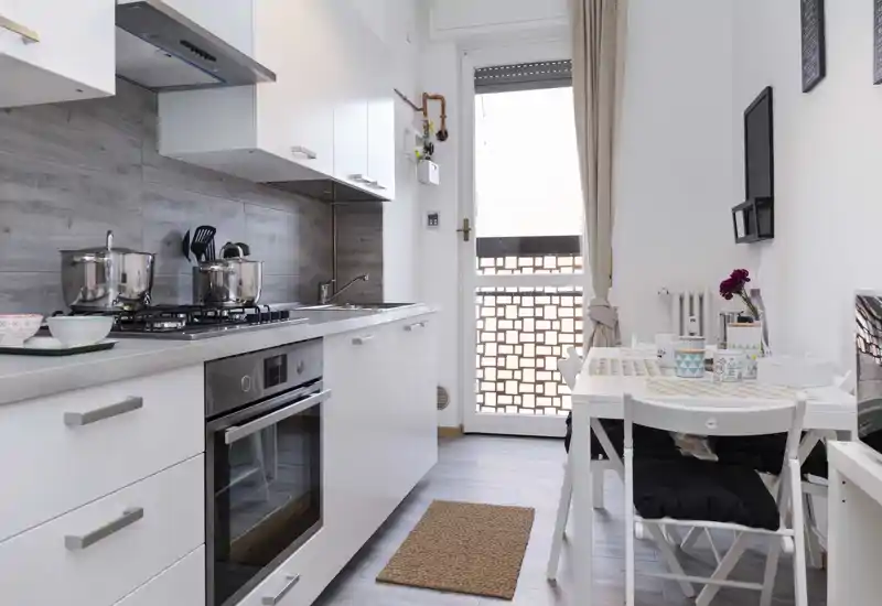 Compact, modern kitchen with a counter and small dining table near a bright window; clean white surfaces and cookware visible.