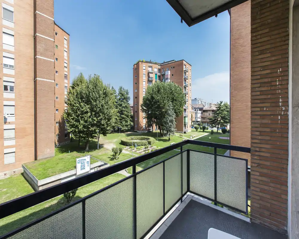 Balcone privato soleggiato con vista su un cortile curato, alberi maturi e palazzi di mattoni vicini.