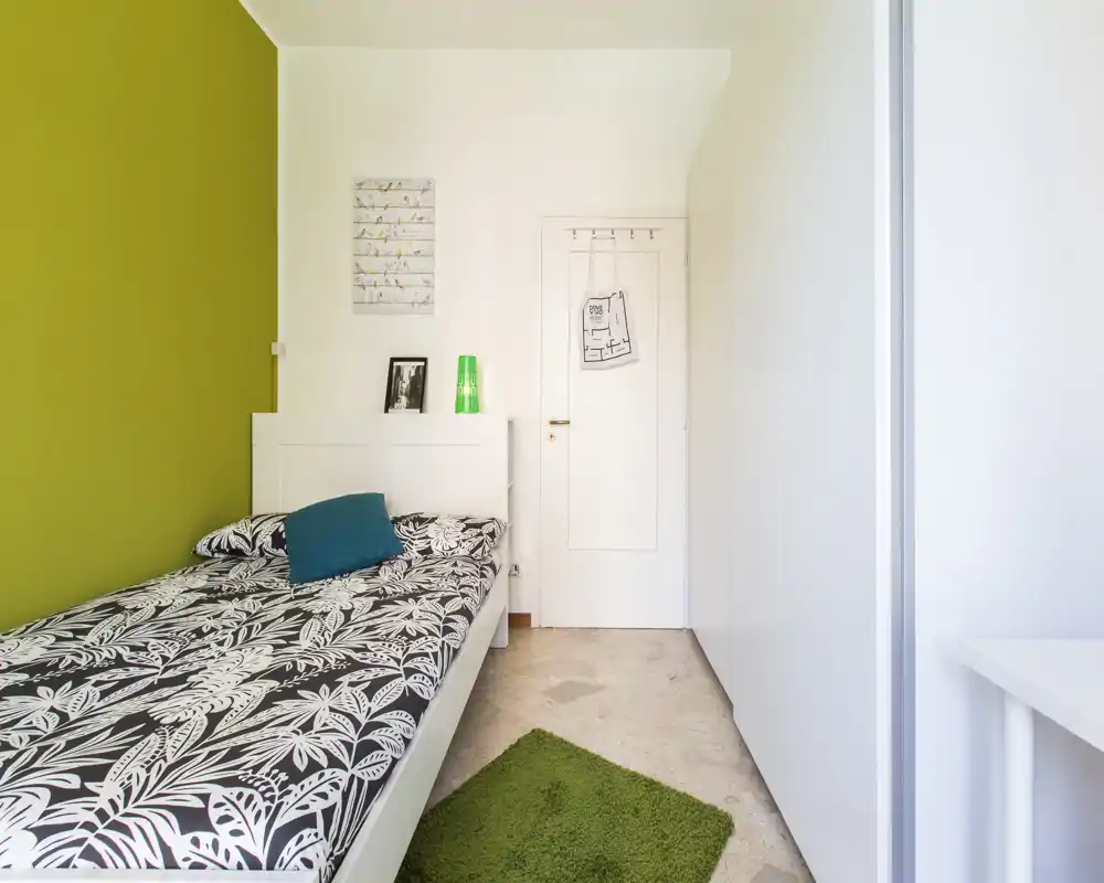 Camera da letto compatta e luminosa con letto singolo contro una parete verde, biancheria con fantasia, piccolo tappeto e armadio — ben illuminata e arredata con gusto.