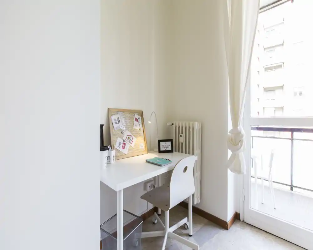 Angolo studio luminoso con scrivania bianca, sedia, lampada e bacheca vicino alla porta del balcone che lascia entrare luce naturale — ideale per studiare o lavorare da casa.