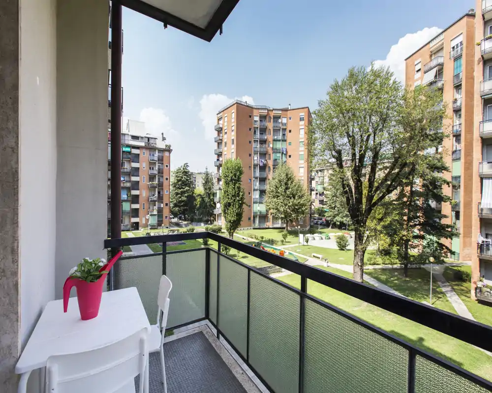 Balcone soleggiato con vista su un cortile alberato e condomini; piccolo tavolo e sedie per un angolo relax.