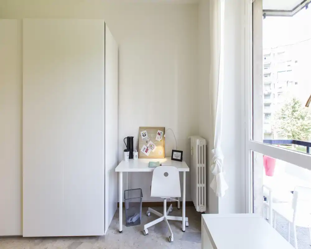 Angolo di camera minimale con un armadio bianco e una piccola scrivania vicino a una grande finestra. Luce naturale abbondante, arredo semplice e una postazione compatta rendono lo spazio pratico e ordinato.