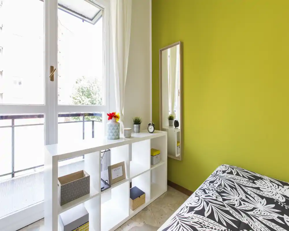 Angolo camera da letto moderno e luminoso con copriletto a fantasia, scaffalatura aperta usata come separé e una grande finestra che dà su un piccolo balcone, con tanta luce naturale.