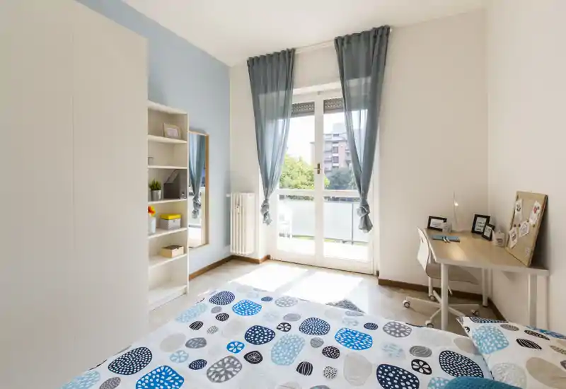 Dormitorio luminoso y ordenado con cama doble con ropa de cama estampada, escritorio junto a la ventana y estantería integrada. Las grandes puertas al balcón dejan entrar mucha luz natural y los tonos azules suaves generan un ambiente tranquilo.