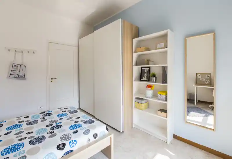 Dormitorio moderno y luminoso con ropa de cama estampada, un gran armario y estantería abierta; la luz natural inunda la estancia y un espejo de cuerpo entero añade profundidad.