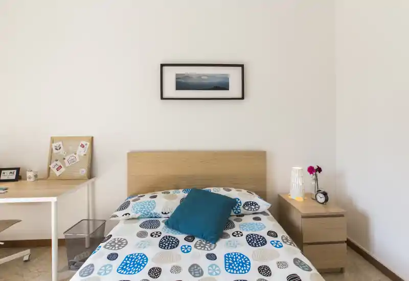Dormitorio luminoso y minimalista con cama doble central, edredón con estampado y cojín turquesa; mesita con lámpara y escritorio a un lado.