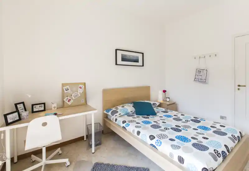 Dormitorio individual luminoso y ordenado con cama de madera, ropa de cama estampada y pequeño escritorio, ideal para estudiantes o inquilinos individuales.