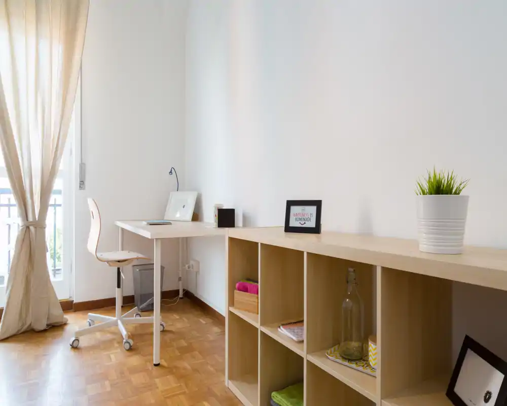 Bright, minimalist home office corner with a desk, swivel chair and open shelving, gently lit by a large window with sheer curtains.