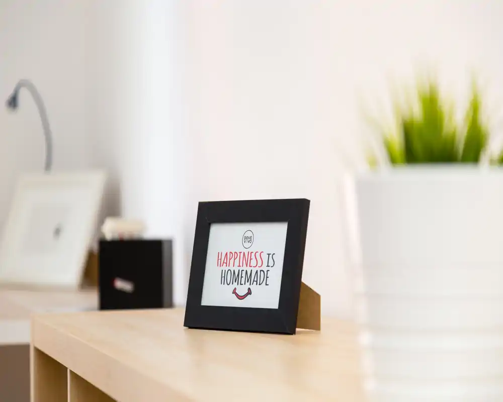 Close-up detail of a wooden tabletop with a small framed sign reading "Happiness is homemade" and a blurred potted plant; this is a decorative detail shot rather than a full room view.
