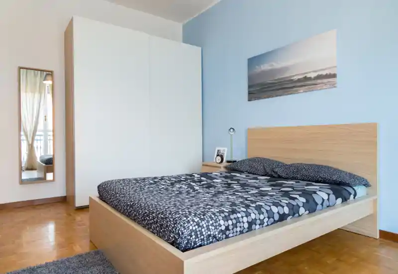 Bright, modern bedroom with a wooden bedframe, patterned bedding, wardrobe and a full-length mirror; soft natural light and calming sea artwork create a welcoming atmosphere.