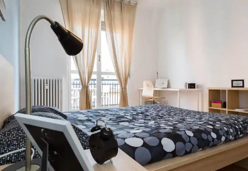 Bright, modern bedroom with a large double bed, patterned duvet and a window letting in abundant natural light. A minimalist desk and shelving provide a functional workspace.