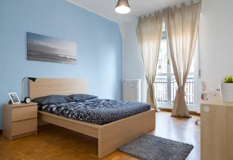 Bright, modern bedroom with a wooden double bed, bedside table and large window letting in natural light. Neutral decor and soft textiles create a calm, welcoming atmosphere.