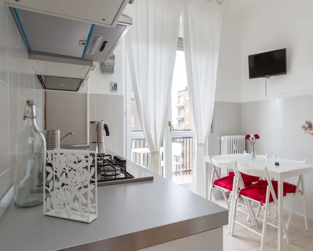 Bright modern kitchen with a gas hob, countertop and a small dining table next to a balcony door; white décor with red cushions adds a cozy touch.