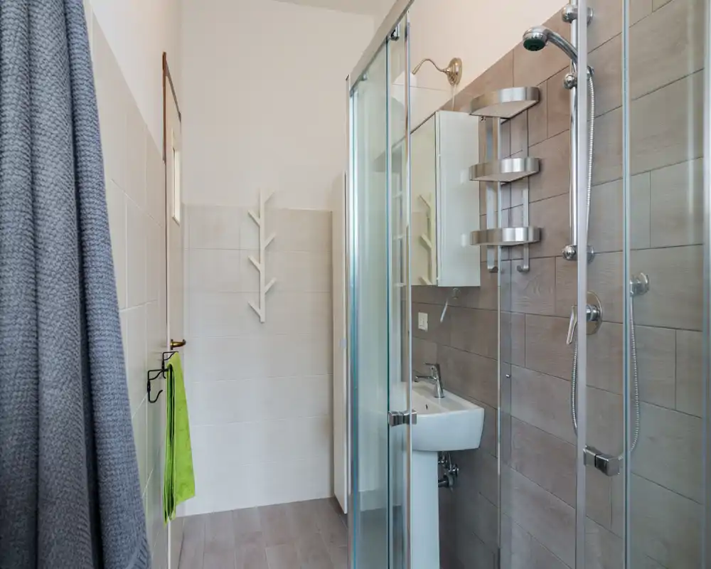 Bright, modern bathroom with a glass shower enclosure, wall-mounted shower head, and a small pedestal sink. Neutral tiles and chrome fixtures create a clean, contemporary look.