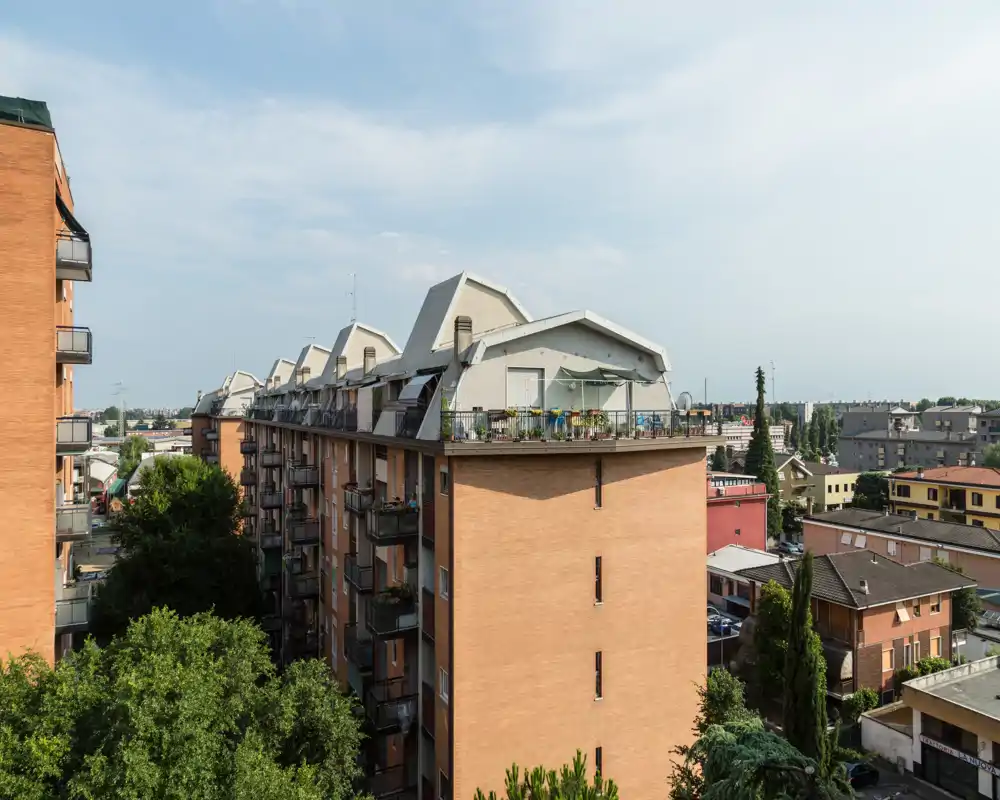 View of residential rooftops and terraces from an upper-floor vantage, showing balconies, rooftop gardens and neighboring buildings under a bright sky.