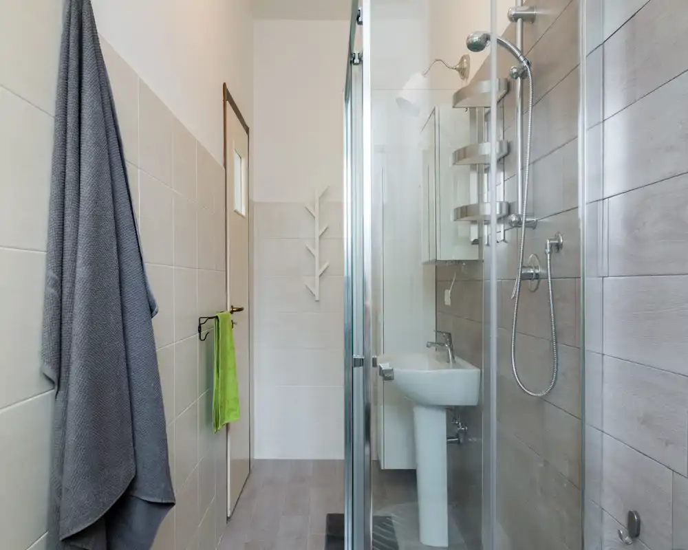 Modern, compact bathroom with a glass-enclosed shower, pedestal sink and neutral tilework, well lit and neatly arranged.