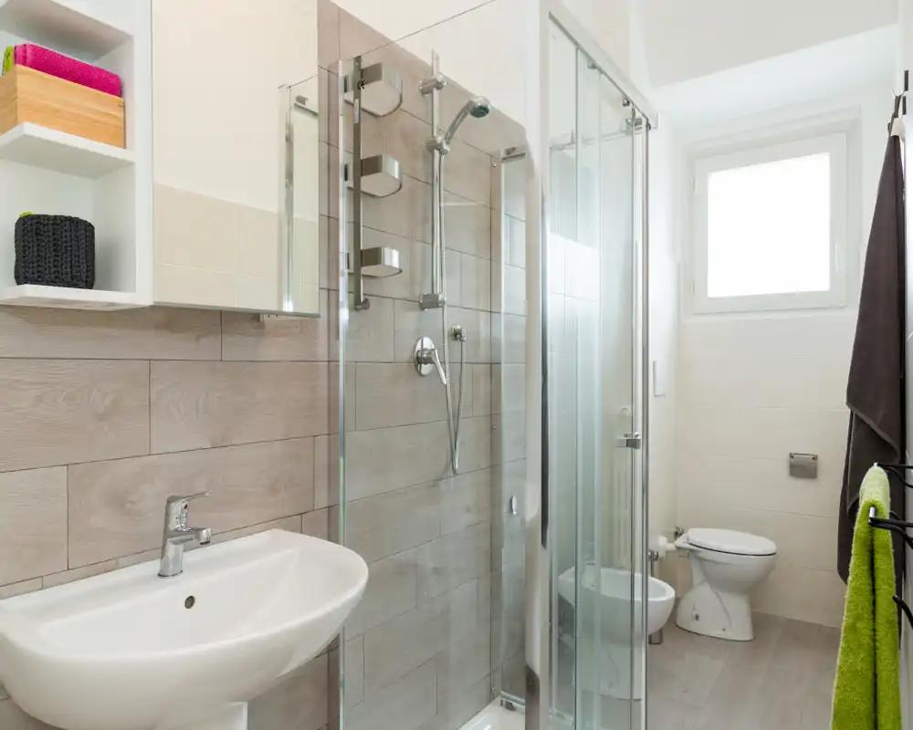 Bright, modern bathroom with glass-enclosed shower, wall-mounted fixtures, sink and toilet. Natural light from a window creates a fresh, clean atmosphere.