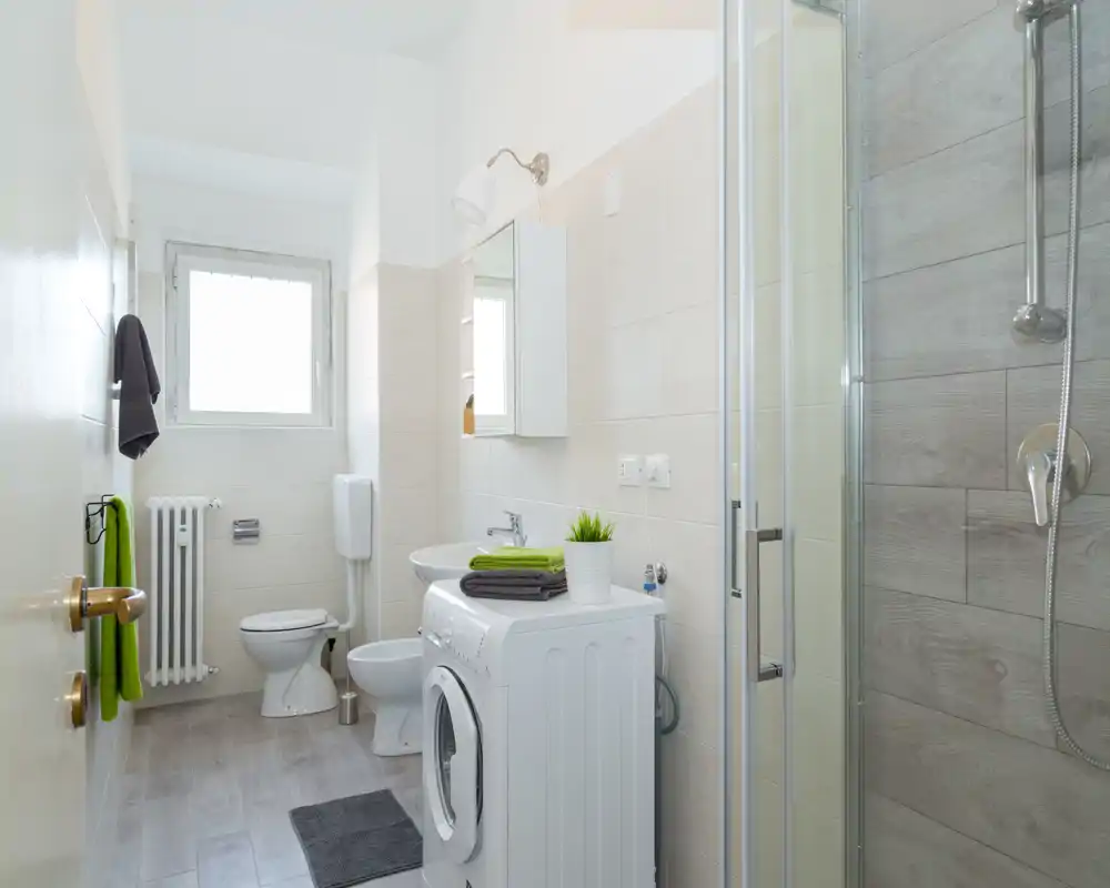 Modern, bright bathroom with a glass shower, toilet, sink, and a front-loading washing machine. Neutral tiles and natural light create a clean, functional space ideal for short-term rentals.