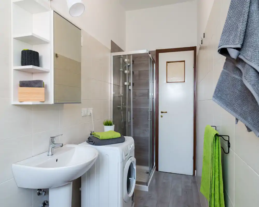 Well-lit modern bathroom with a corner shower, sink, washing machine and tidy towel storage; neutral tiles and green accents give a fresh, clean look.