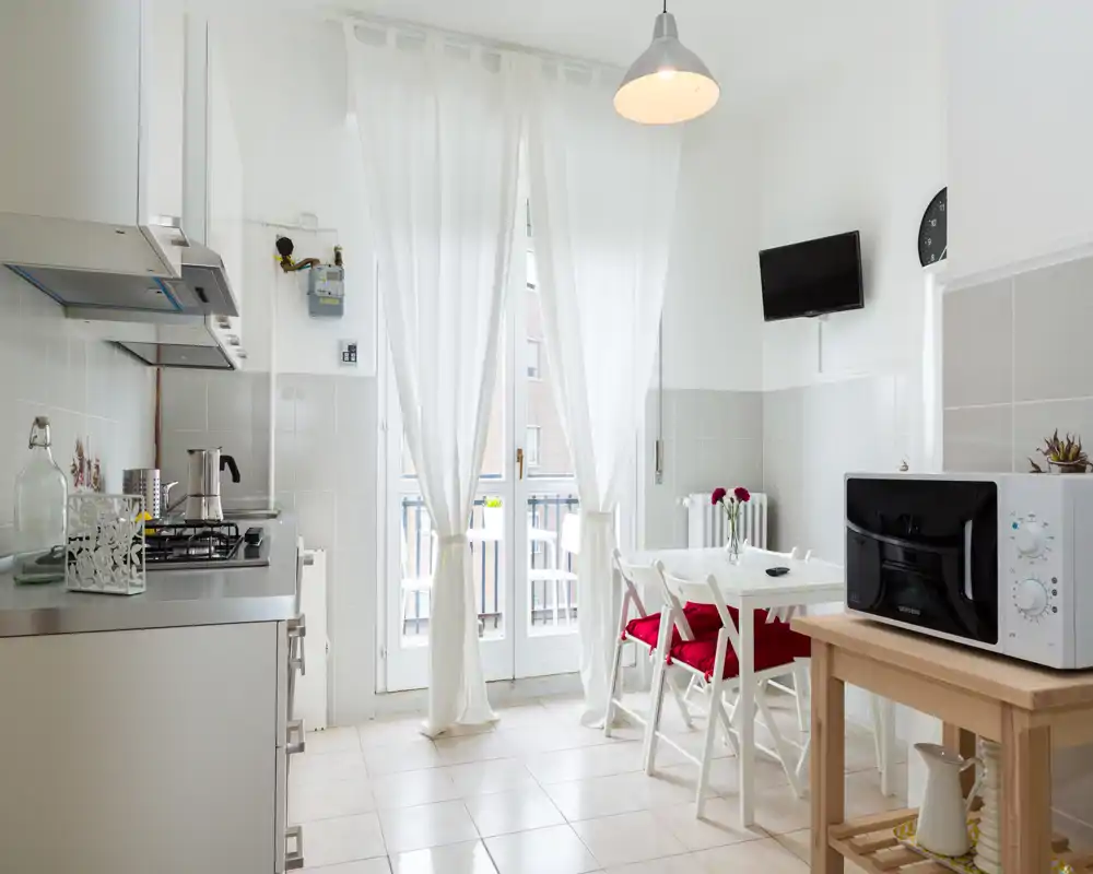 Bright, compact kitchen with a small dining table and chairs, stainless-steel counters, microwave and large glass doors opening to a balcony; modern, airy and well-lit space.